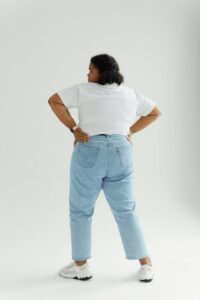 Rear view of a confident plus size woman in denim pants and white shirt standing on a white background, embracing body positivity.