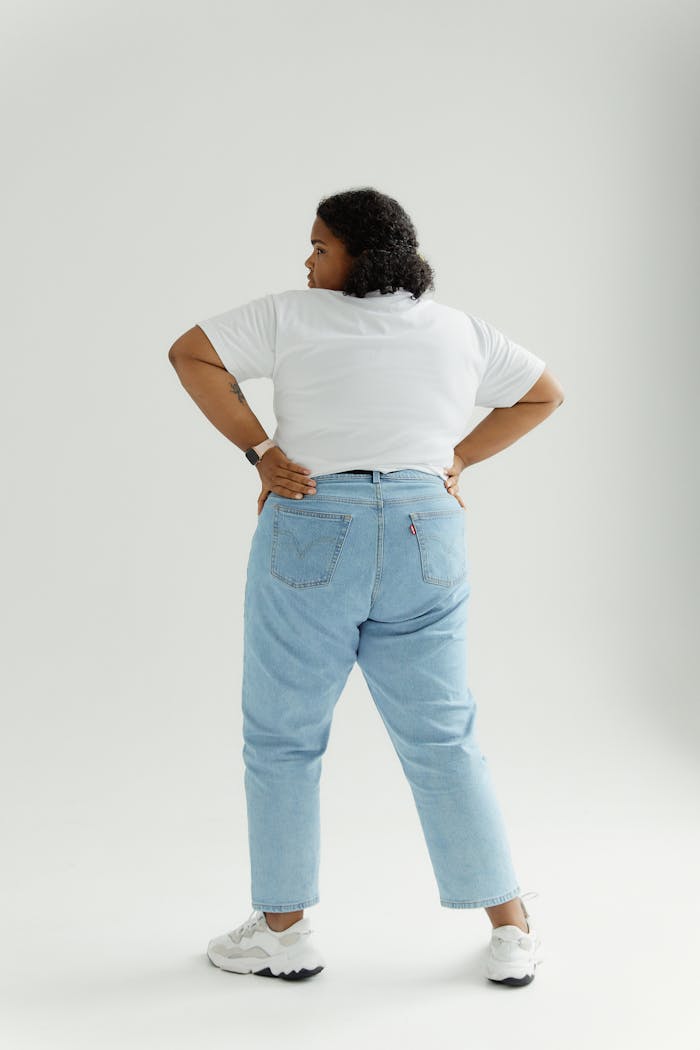 Rear view of a confident plus size woman in denim pants and white shirt standing on a white background, embracing body positivity.